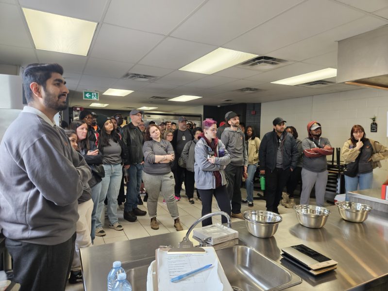A large group listens to a speaker beside stainless-steel counters in a commercial kitchen.