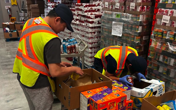 Two volunteers in high-visibility vests sort packaged food into boxes in a warehouse.
