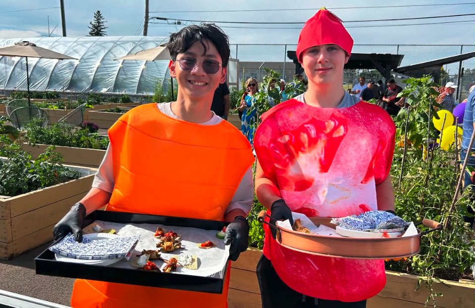 Two young volunteers in colourful food costumes carry serving trays in a community garden.