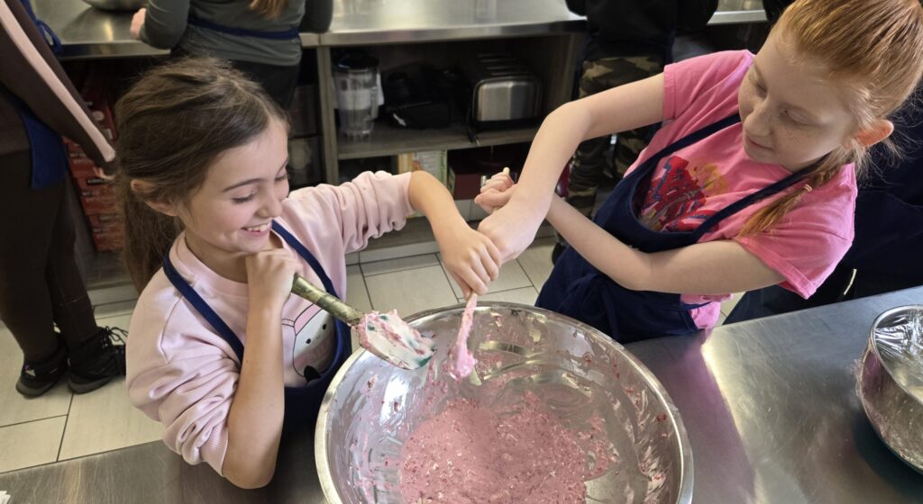 Two children wearing aprons stir a pink mixture in a large stainless-steel bowl.