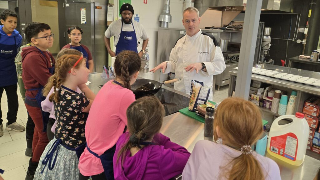A chef demonstrates cooking to children gathered around a commercial kitchen counter.