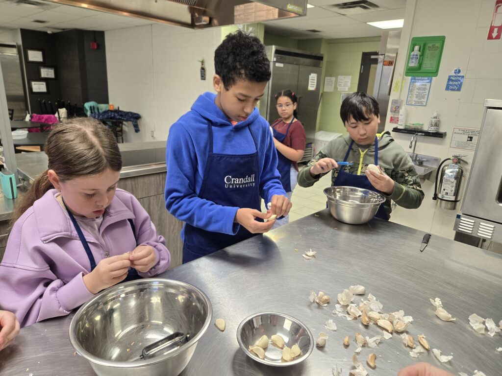 Children wearing aprons peel garlic at a stainless-steel kitchen counter.
