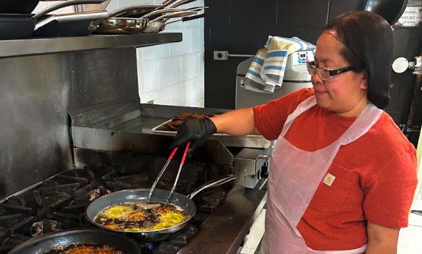 A cook uses tongs to prepare food in a frying pan on a commercial stove.