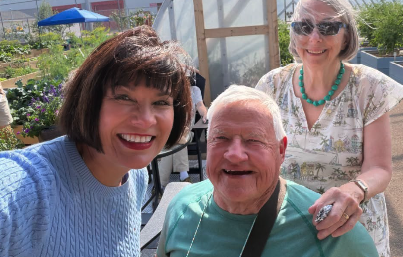 Three adults smile for a photo in a sunny community garden.